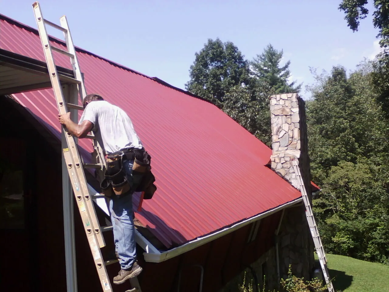 Roofer on a ladder beside a red metal roof and stone chimney — Soffit & Fascia Repair by Roofing Brothers in East Bridgewater